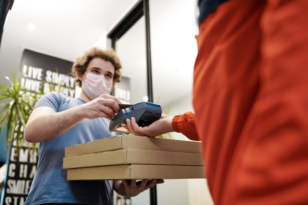 A man wearing a face mask holding pizza boxes using a card reader machine held by another person
